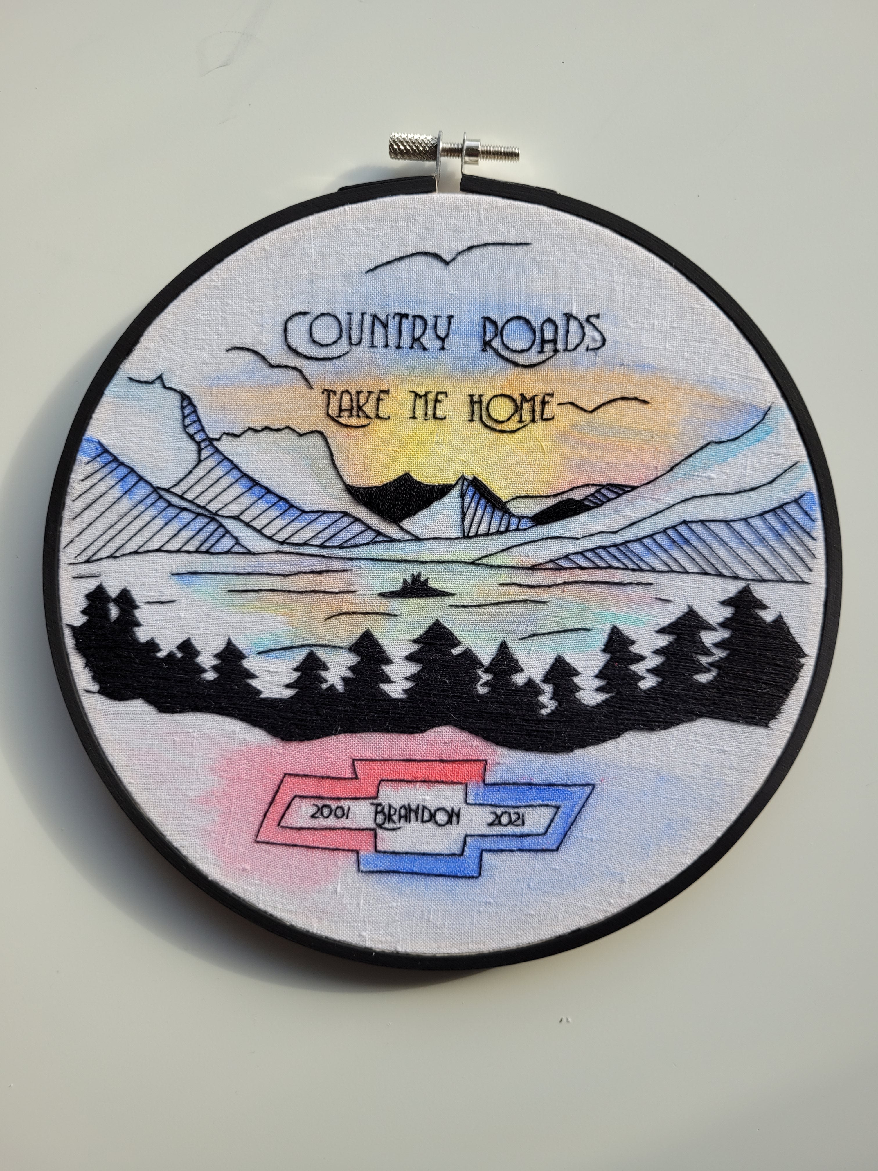 embroidery hoop of mountains and valley. text: 'Country Roads Take me Home 2001 Brandon 2021"