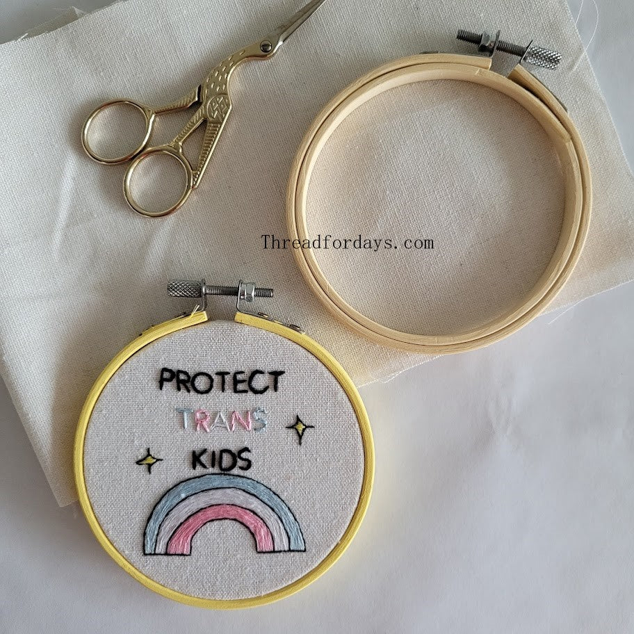 protect trans kids hoop on aida fabric. another 4 inch embroidery hoop and scissors in the photo