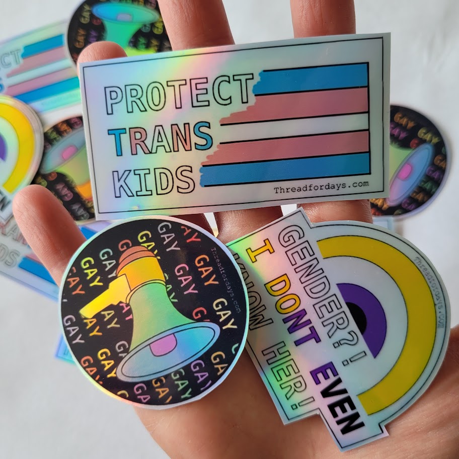 three stickers (protect trans kids, say gay, gender i dont even know her) held in hand