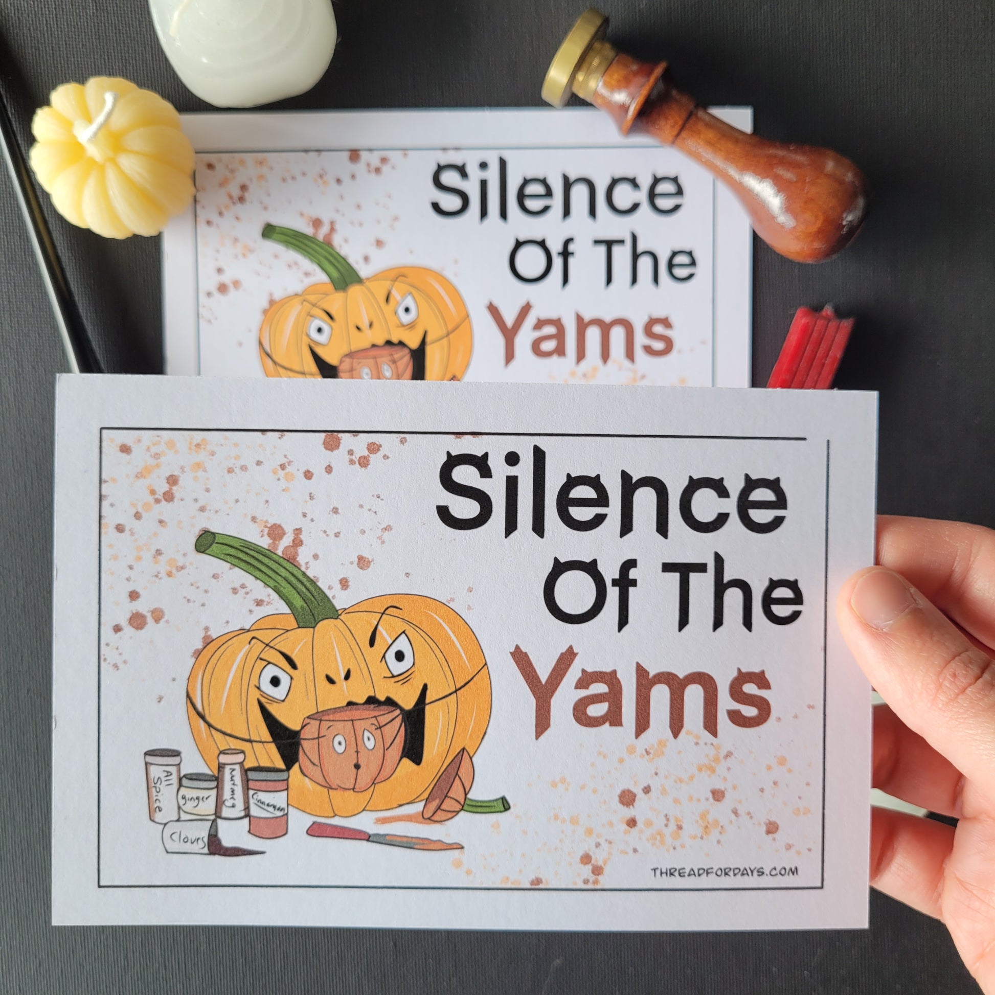 front of silence of the yams postcard featuring a pumpkin about to eat a smaller pumpkin. the top of the pumpkin has been cut off with a knife and pumpkin spice seasoning jars surround the large pumpkin