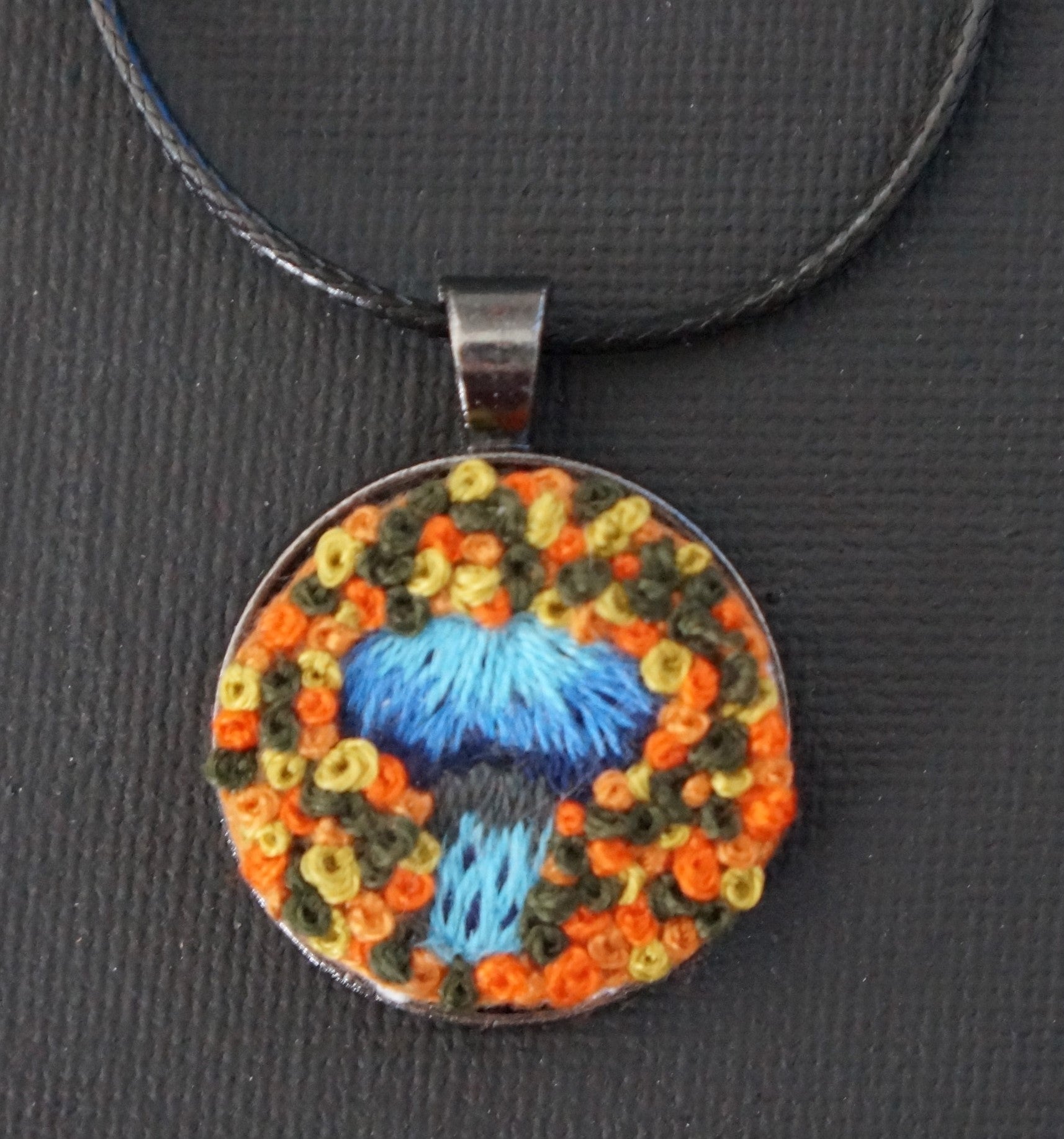 a bright blue embroidered mushroom with an orange and blue french knot textured background. Set in a wearable pendant